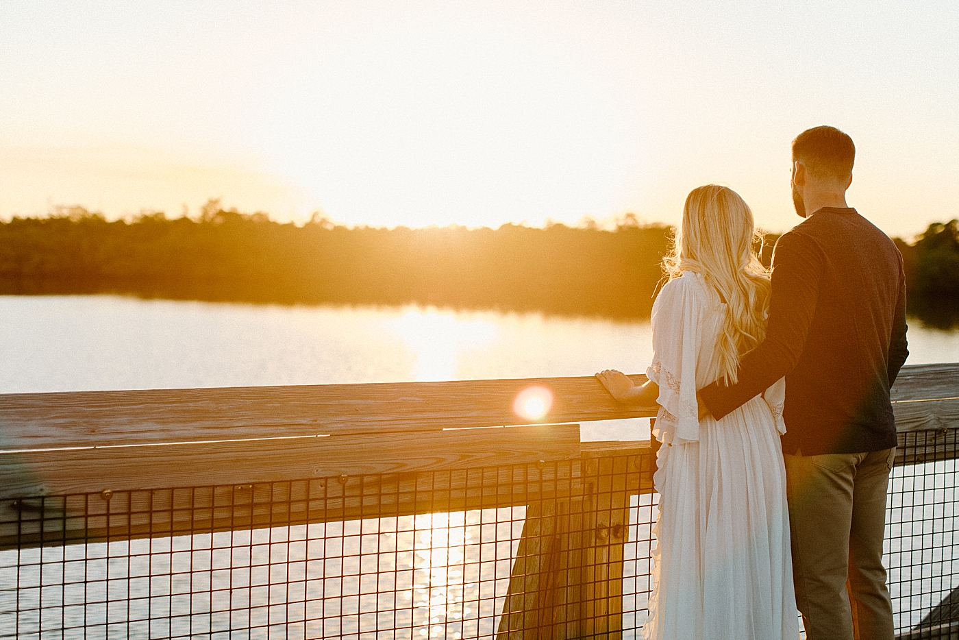 Couple with arms around each other as they watch the sunset on calm river Jupiter Engagement Photography captured by South Florida Engagement Photographer Erika Tuesta Photography