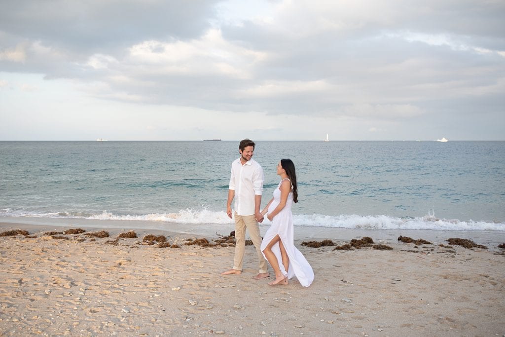 Pregnant couple holding hands walking along the beach shoreline