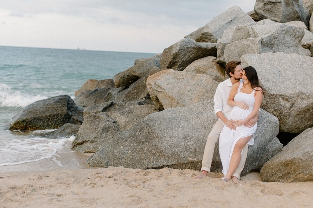Husband kissing pregnant wife sitting on beach rocks