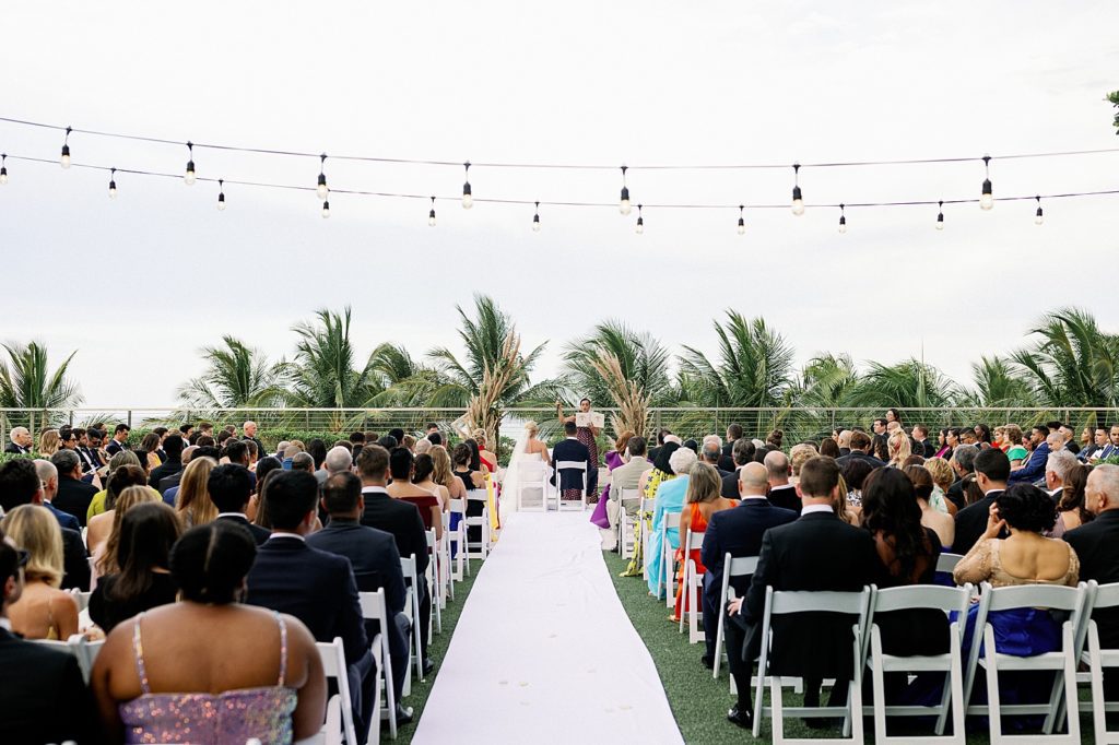 Wide shot of outdoor Ceremony with hanging lights and bride and groom sitting in front of officiant