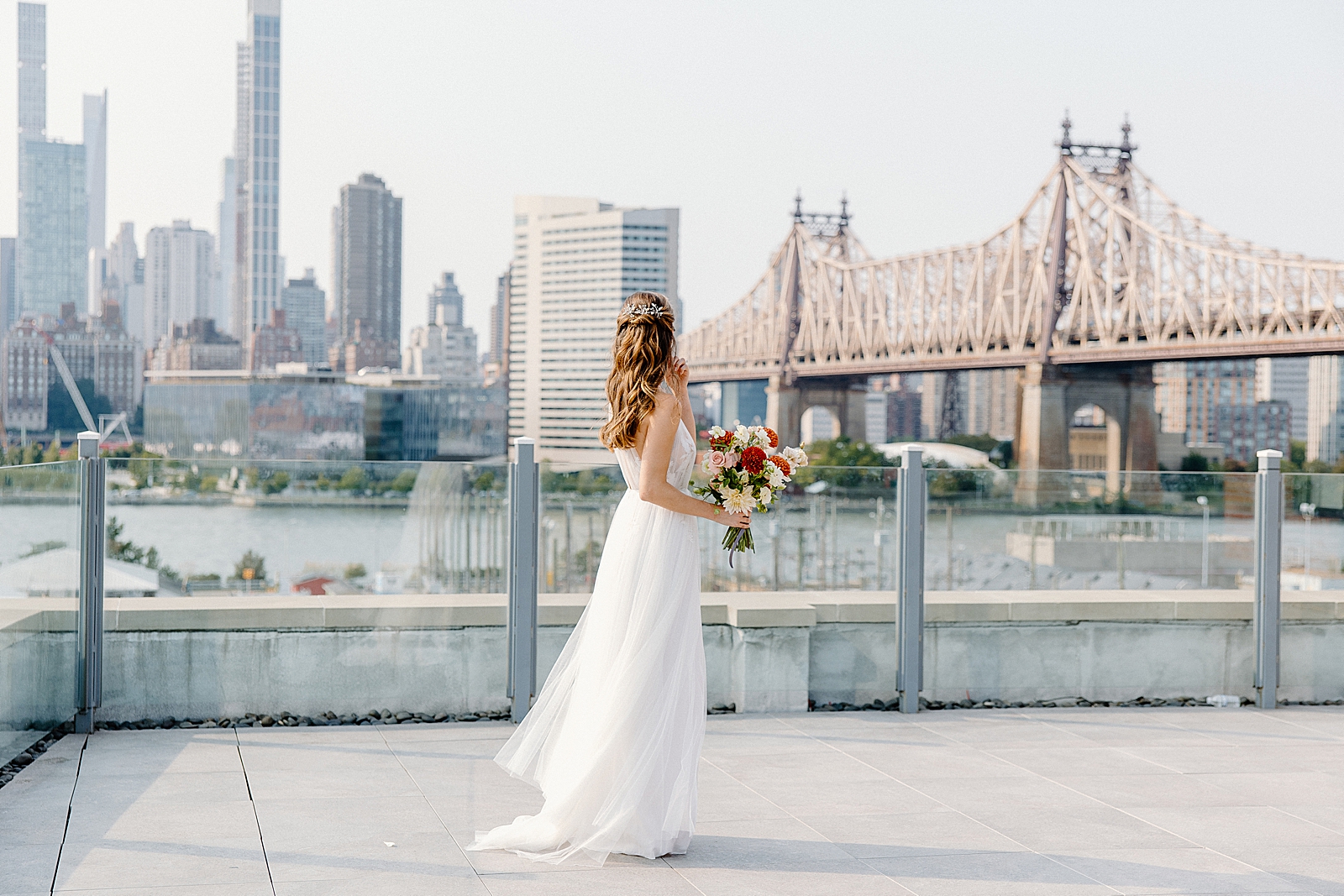 Bride holding bouquet and looking across East River at Manhattan