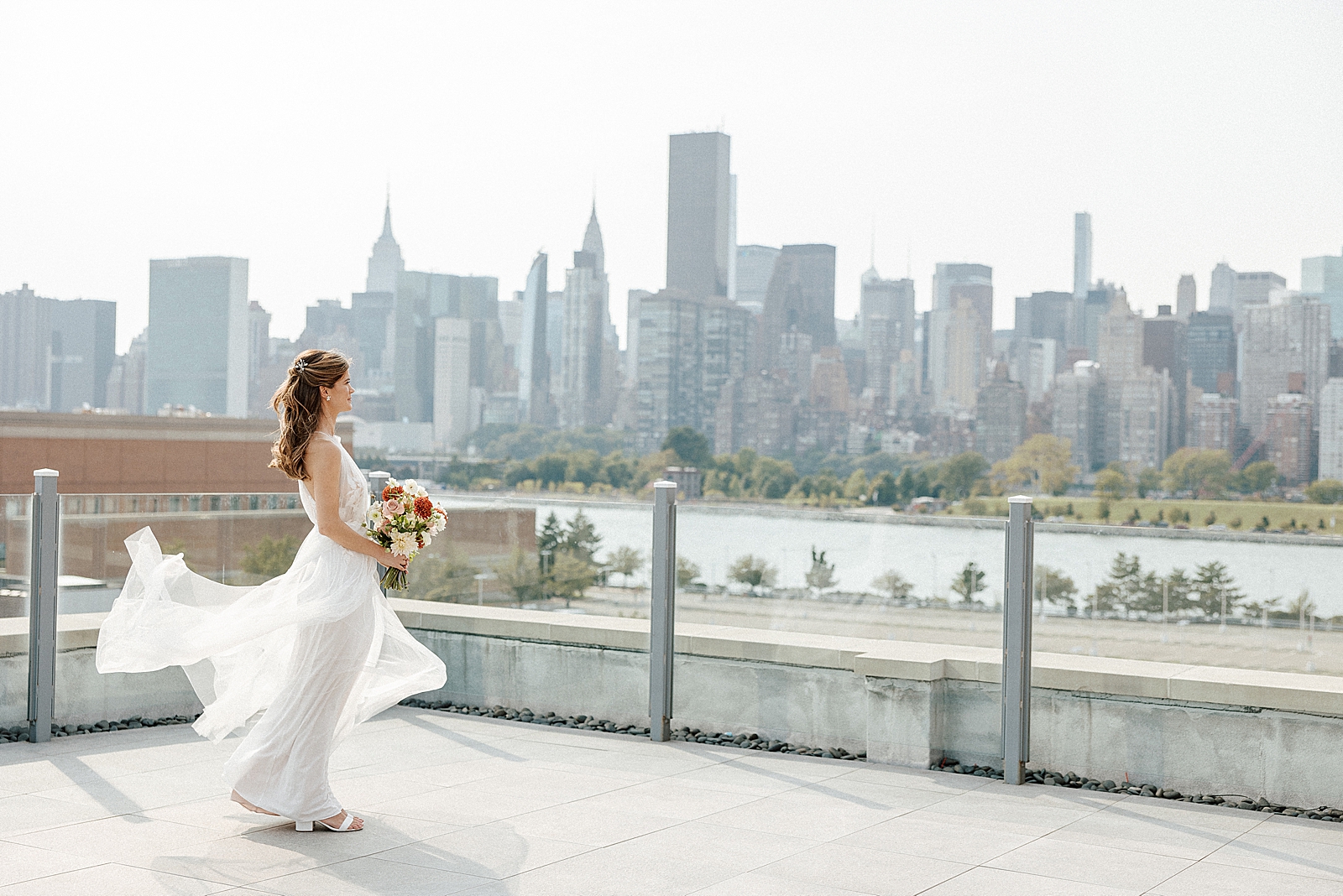 Bride holding bouquet and looking across East River at Manhattan with Empire state building and Chrysler building view